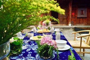 Ein Essbereich im Freien mit einem für eine Mahlzeit gedecktem Tisch, umgeben von Holzmöbeln und einem traditionellen Gebäude im Hintergrund.