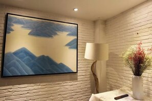 Una habitación con un cuadro grande enmarcados de montañas, una lámpara blanca y un jarrón con flores.