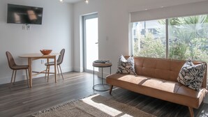 A modern living room with a sofa, a dining table with chairs, a TV mounted on the wall, and a large window with a view of greenery. - Agave on the Beach (Miami Beach)