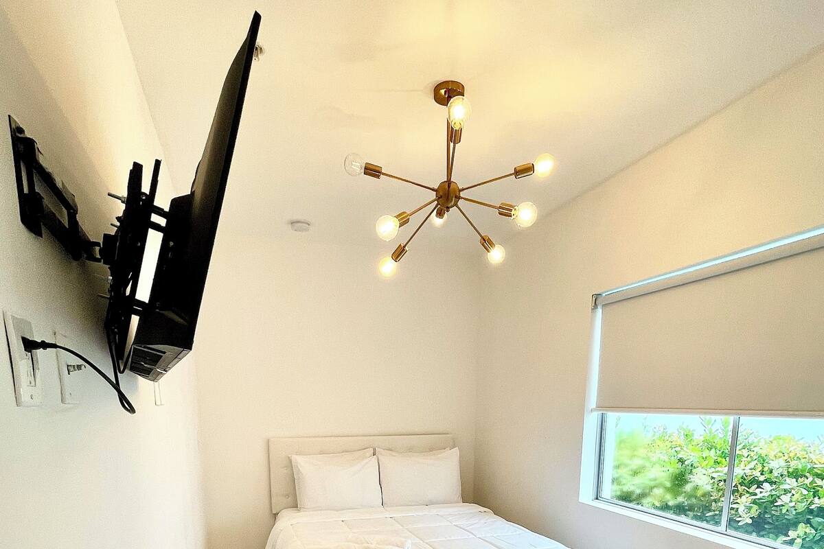 A bedroom with a bed, a round table, a TV mounted on the wall, a ceiling light fixture, and a window with a view of greenery.