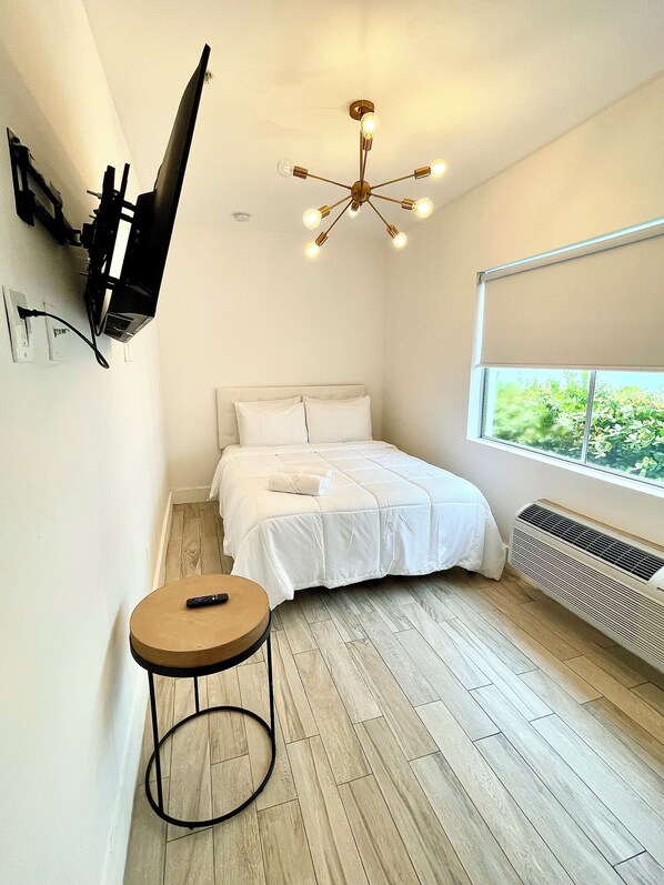 A bedroom with a bed, a round table, a TV mounted on the wall, a ceiling light fixture, and a window with a view of greenery. - Agave on the Beach (Miami Beach)