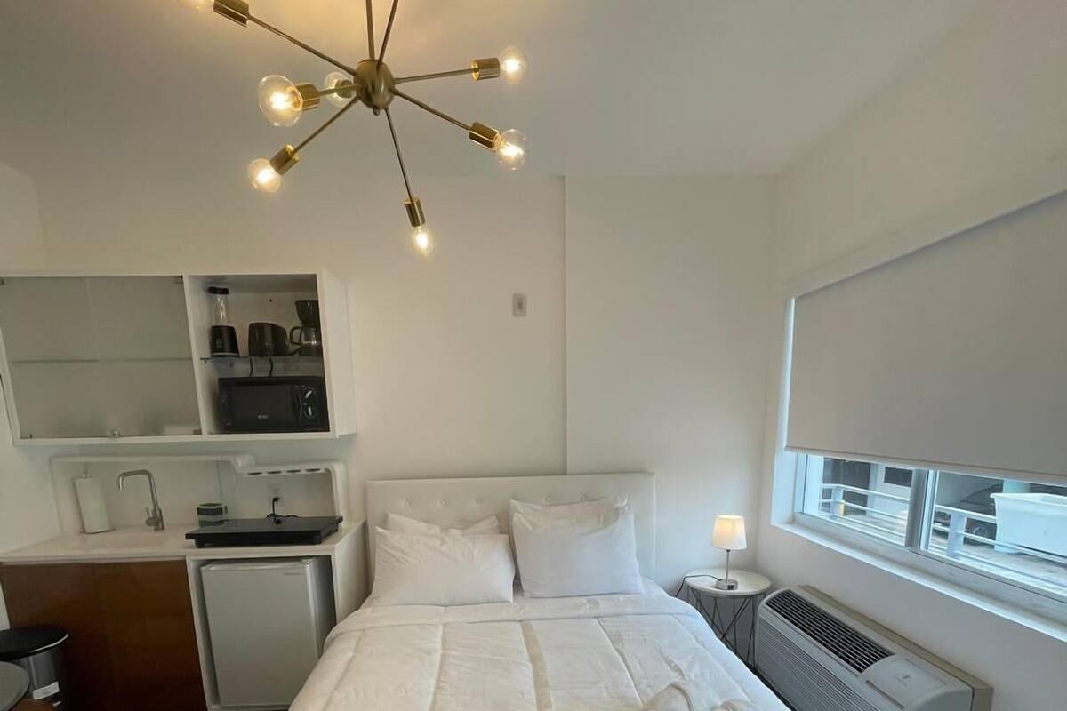 A modern hotel room with a central bed, a built-in kitchenette, a window with a view, and a unique chandelier.