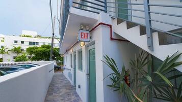 Caminho estreito que leva a uma placa de saída num edifício, com uma escadaria e área de piscina visíveis à esquerda.