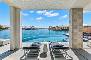 A poolside area with lounge chairs, a table with two glasses of wine, and a view of a marina with boats and buildings. - Infinity; Lakefront 5-Bedroom with Infinity Pool, Pet Friendly, & Fun Filled!
 (Indio)