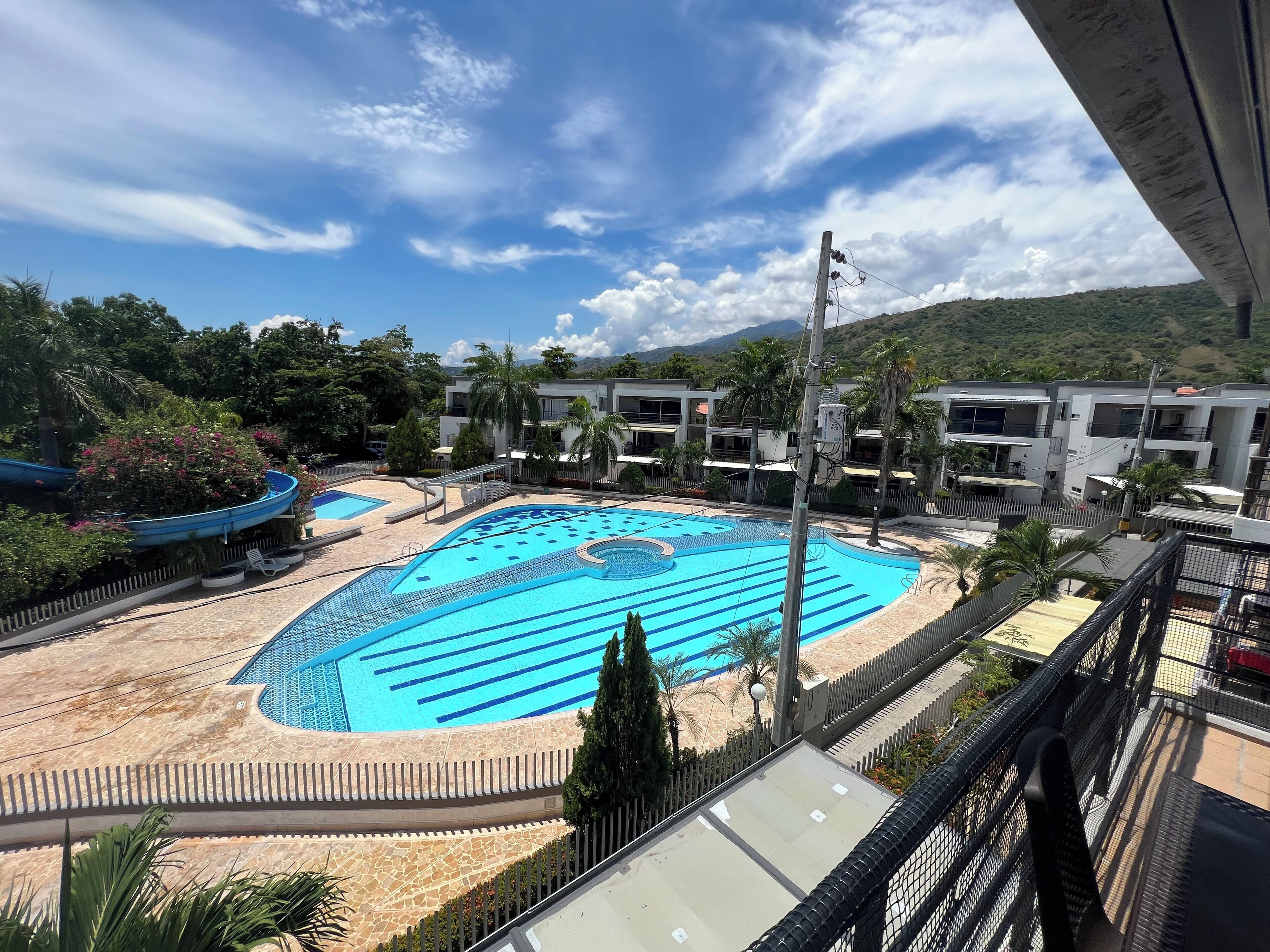 A large outdoor swimming pool with a blue and white design, surrounded by palm trees and a mountainous backdrop.