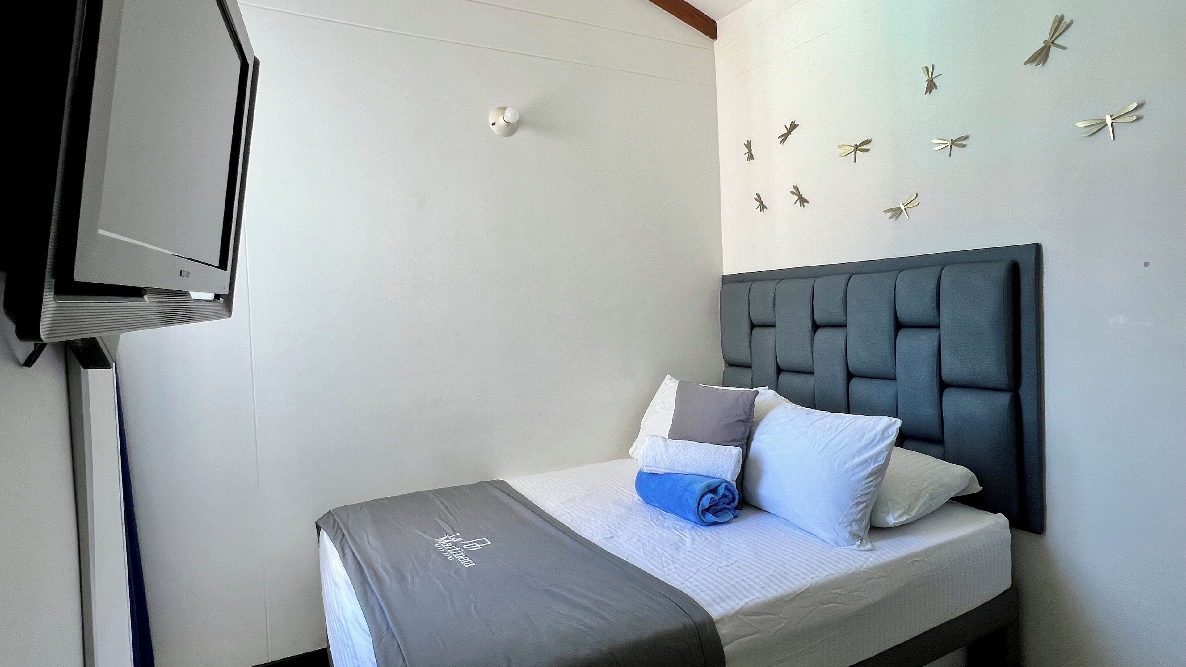 A neatly made bed with a grey headboard, a blue blanket, and several pillows. A flat-screen TV is mounted on the wall to the left.