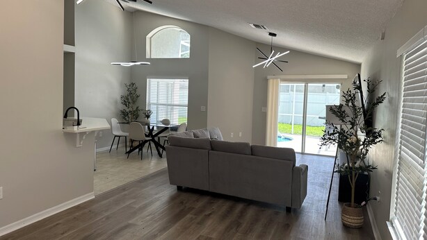 A modern living room with a gray sofa, dining table, and chairs. There are two large windows with blinds, a ceiling fan, and a potted plant.