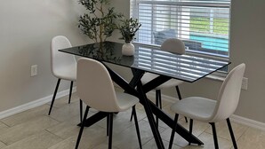 A dining area with a glass table, four chairs, a potted plant, and a modern pendant light.