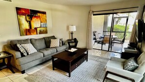 A living room with a grey sofa, a coffee table, and a TV. There's a painting on the wall and a window with curtains.