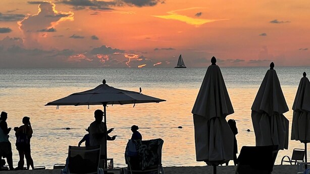 A beach scene with umbrellas and lounge chairs, a sailboat, and a sunset.