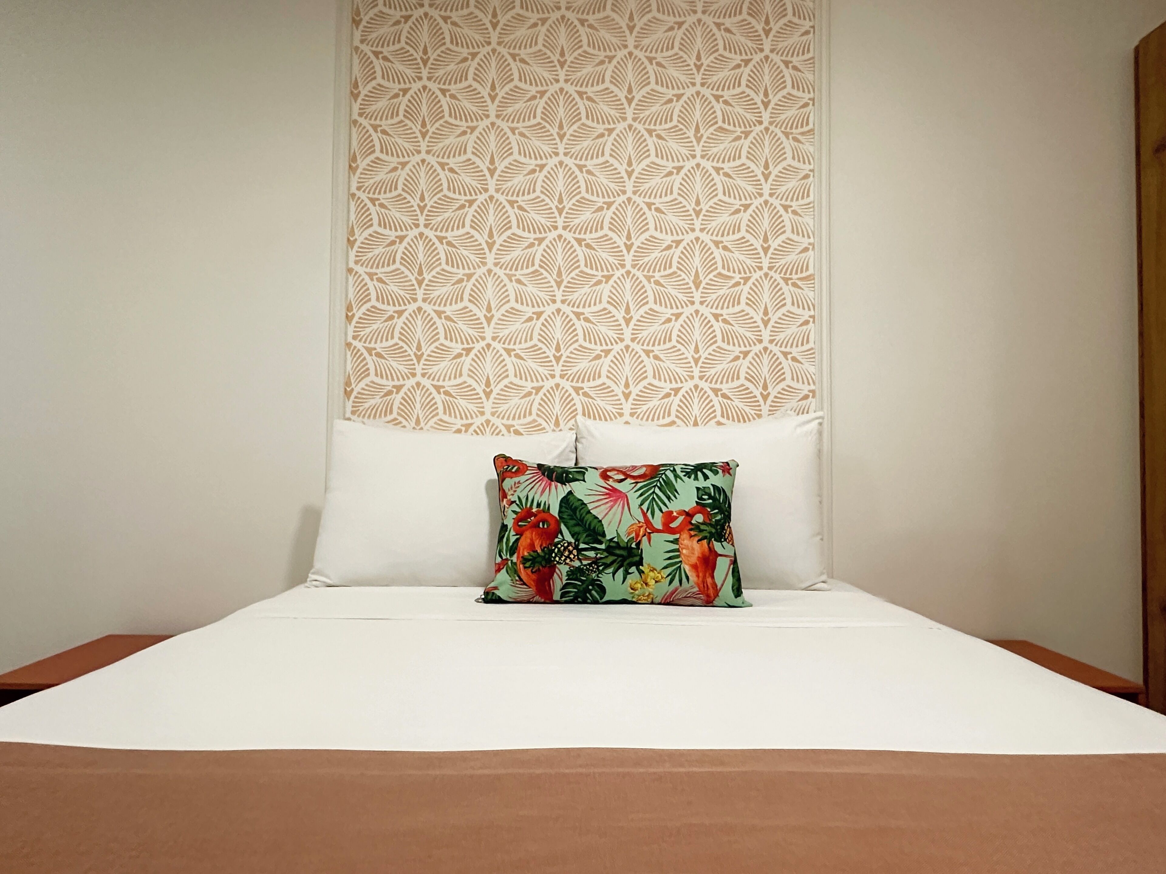 Un lit avec du linge de lit blanc, une tête de lit décorative ornée d’un motif de feuilles et un coussin coloré.