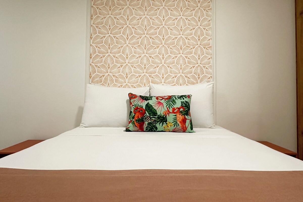 A bed with white linens, a decorative headboard with a leaf pattern, and a colorful pillow.