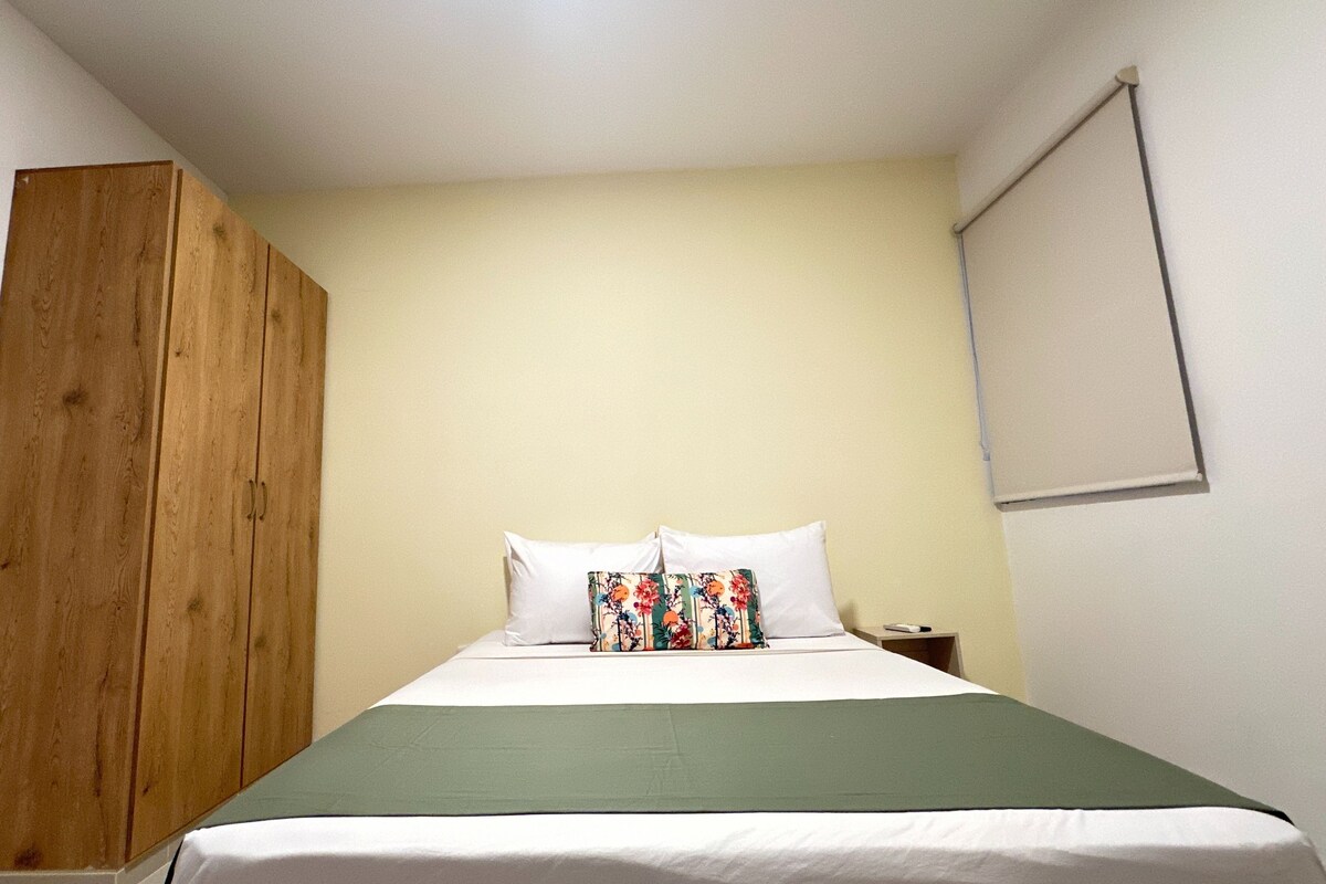 A bedroom with a bed, a wooden wardrobe, a nightstand, and a window with blinds.
