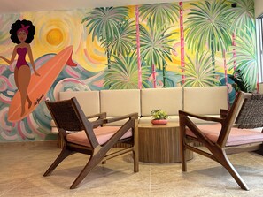Een kamer met een bank, twee houten stoelen, een bijzettafeltje met een plant en een kleurrijke muurschildering met tropisch thema.