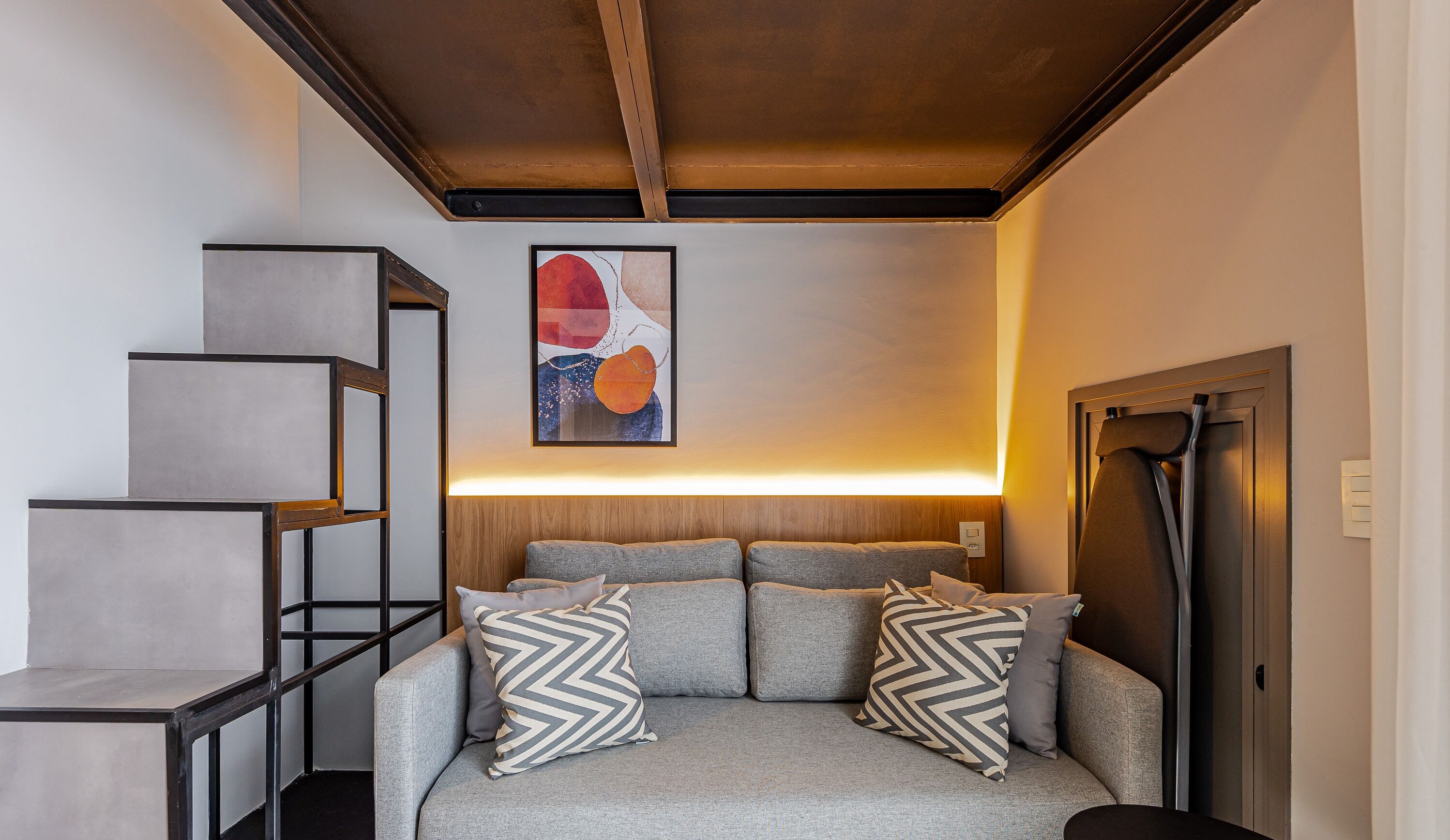 A modern living room with a grey sofa, two patterned pillows, a wooden headboard, and a staircase with a metal railing.