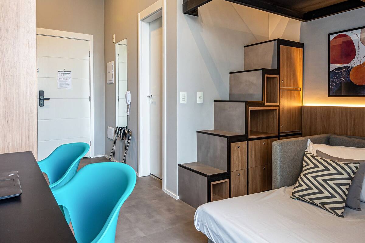 A modern hotel room with a bed, a desk, and a chair. There is a staircase leading to an upper level.