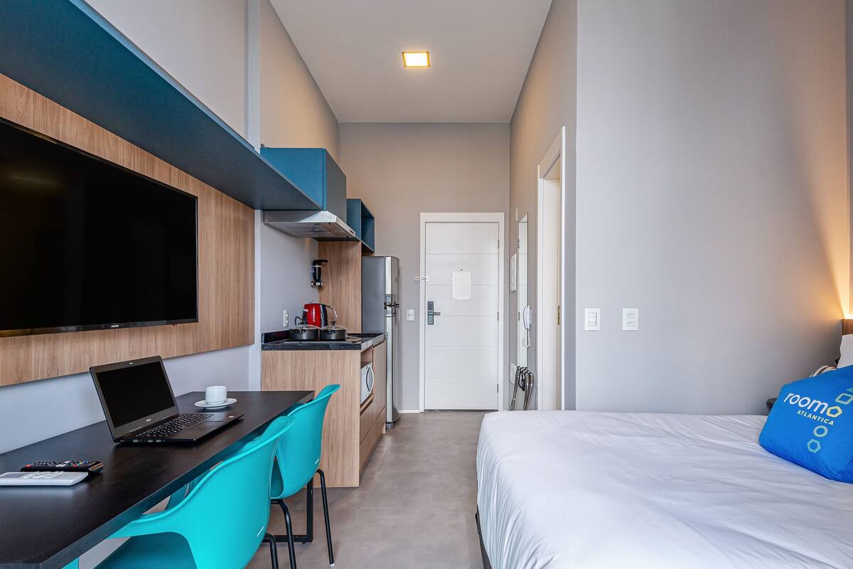 A compact hotel room with a bed, a desk with a laptop, a TV, and a kitchenette.