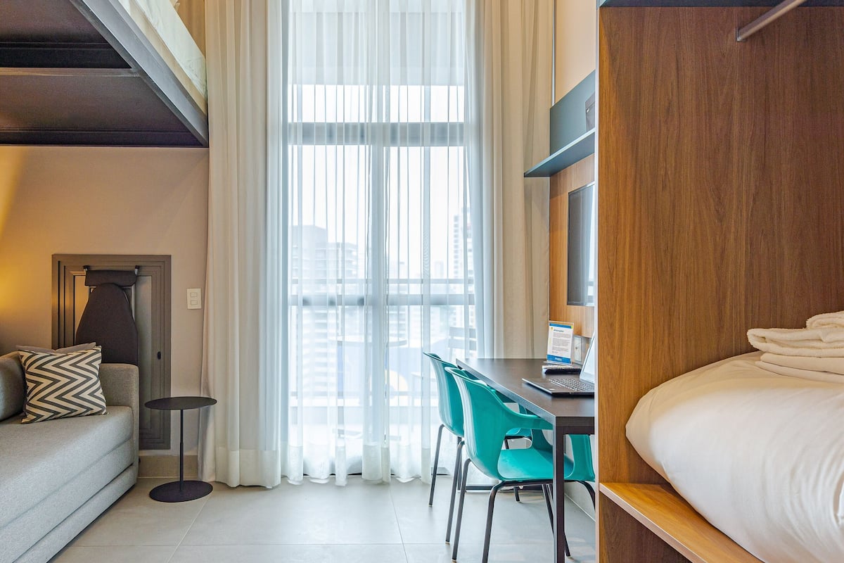 A hotel room with a bed, a desk with a laptop, a chair, a sofa, and a window with curtains.