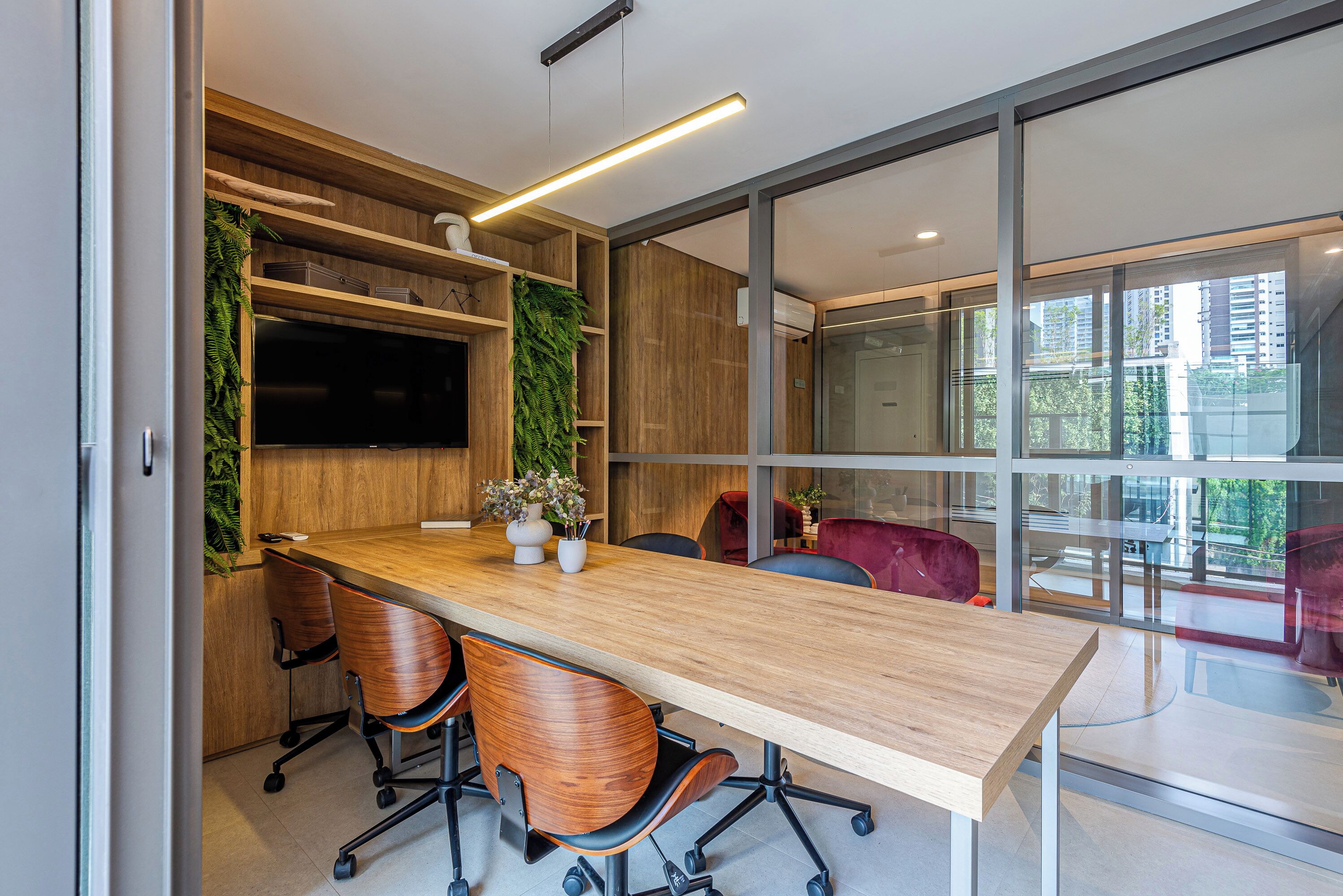 A modern conference room with a long wooden table, four office chairs, a flat-screen TV, wooden shelving, and a large window offering a view of the city.