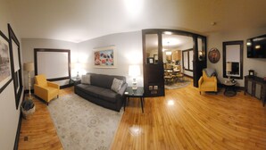 Un salon comprenant un canapé, des fauteuils, une table basse et un tapis. On y trouve également un tableau au mur, une lampe et un téléviseur.
