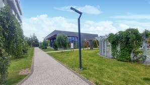 A paved pathway leading to a modern building with a glass facade, surrounded by greenery and a metal fence.