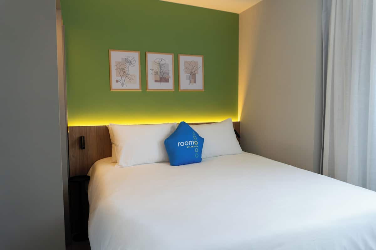 A hotel room with a green accent wall, a bed with white bedding, a blue pillow with the brand name, and three framed floral artworks above the bed.