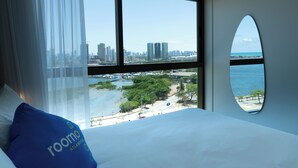 A hotel room with a large bed, a view of a cityscape, and a beach. - Roomo Transamerica Recife Parque do Cais (Recife)