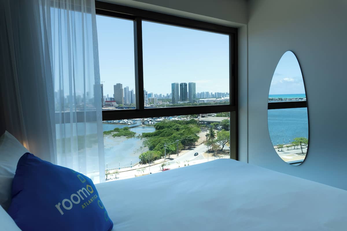 A hotel room with a large bed, a view of a cityscape, and a beach.