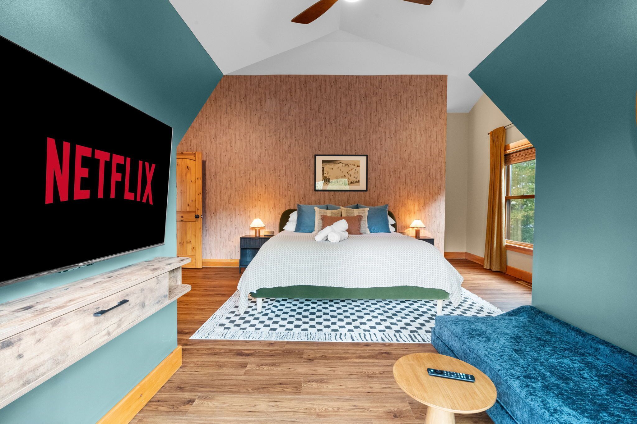 Ein modernes Schlafzimmer mit einem großen Flachbildfernseher, Holzboden, einem Bett mit gemusterten Bettwäsche, einem blauen Sofa und einem kleinen Tisch.
