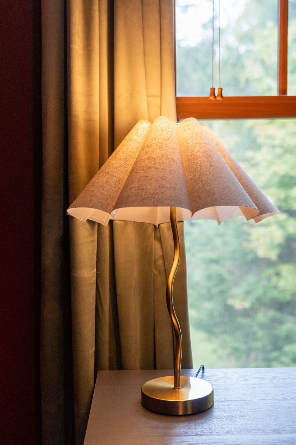 A table lamp with a conical shade on a wooden table, positioned near a window with curtains.
