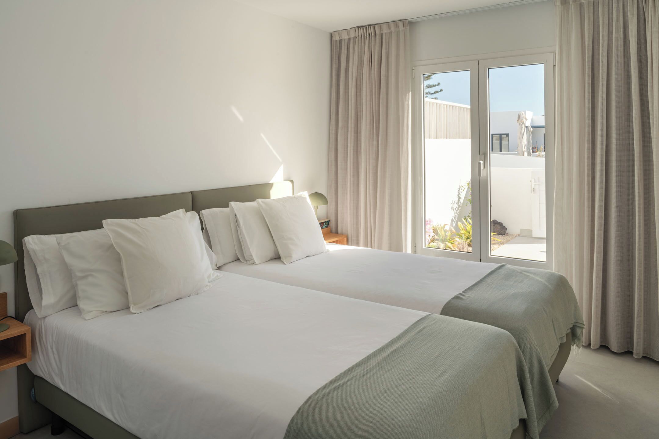 Una cama doble con ropa de cama blanca y un plaid verde, una mesita de noche con una lámpara y un ventanal corredero a un balcón.