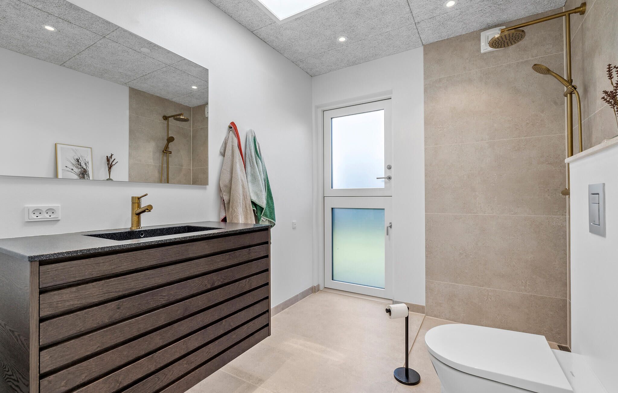 Une salle de bain moderne avec un grand miroir, un lavabo doté d’un plan-vasque foncé, des toilettes et une douche avec une pomme de douche dorée.
