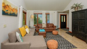 Ein Wohnzimmer mit einem braunen Sofa, einem braunen Ledersessel, einem blauen Hocker und einer schwarzen Kommode. An der Wand hängt ein Gemälde von Bergen, ein Fenster mit Vorhängen und eine Tür mit Fußmatte.
