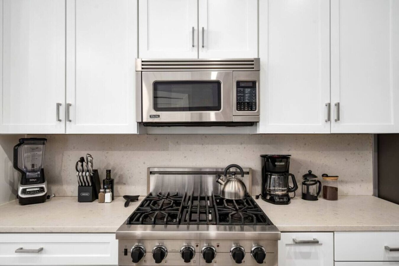A kitchen with white cabinets, a stainless steel microwave, a gas stove, a blender, a kettle, and a coffee maker.