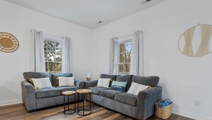 Un salón con dos sofás grises, una mesa de centro y una mesita auxiliar. Hay dos ventanas con cortinas blancas, un reloj de pared y una cesta tejida en el suelo.