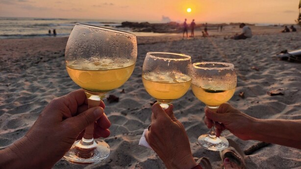 Drei Gläser mit einem Getränk an einem sandigen Strand, im Hintergrund geht die Sonne unter.