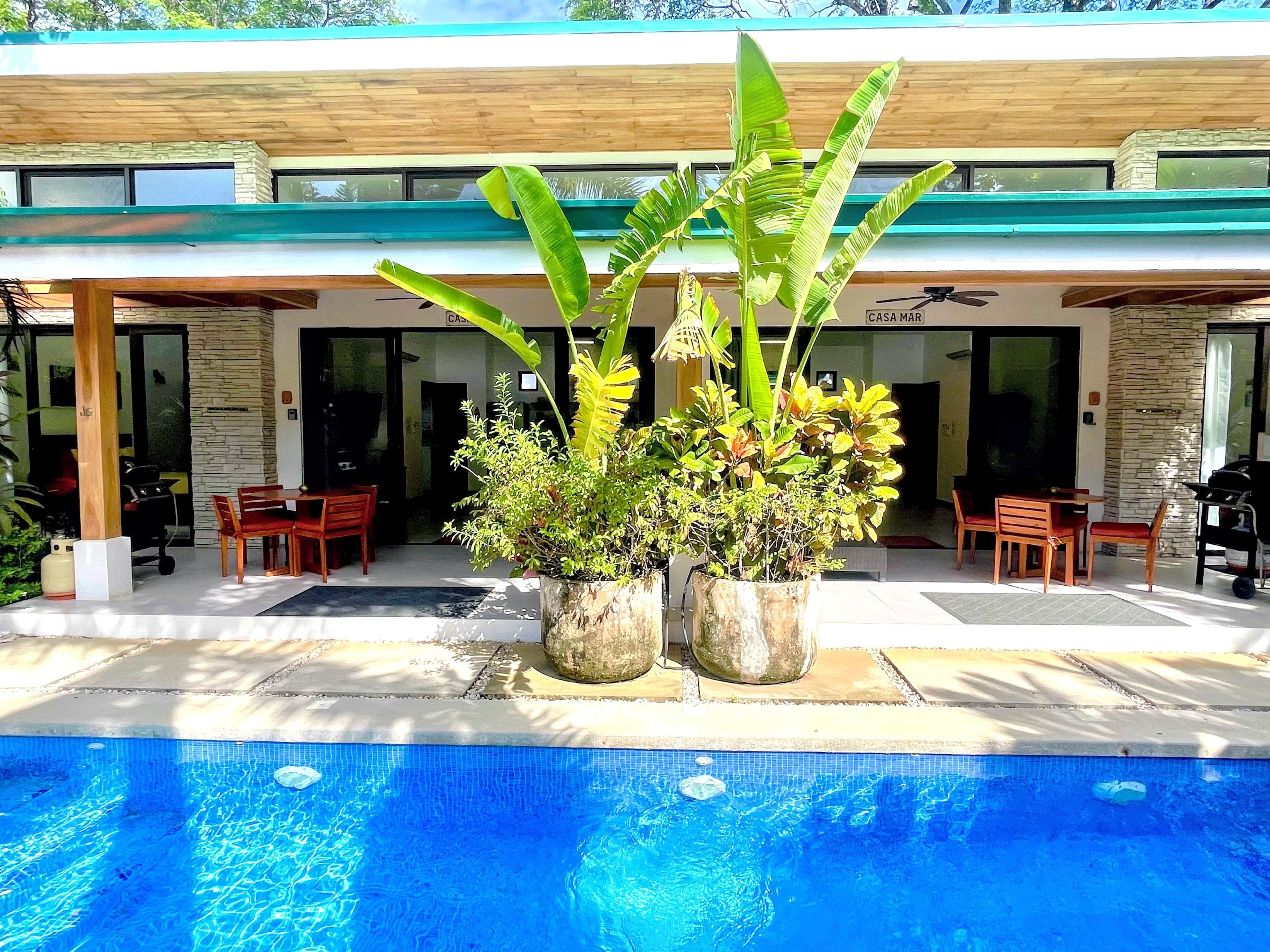 A pool area with a modern house in the background, outdoor seating, and potted plants.