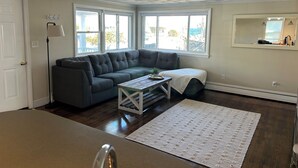 A room with a gray sectional sofa, a small table, a white rug, and a large window with a view of the outdoors. - Charming 3-bedroom house with ocean views, WiFi, AC in delightful Hull (Hull)
