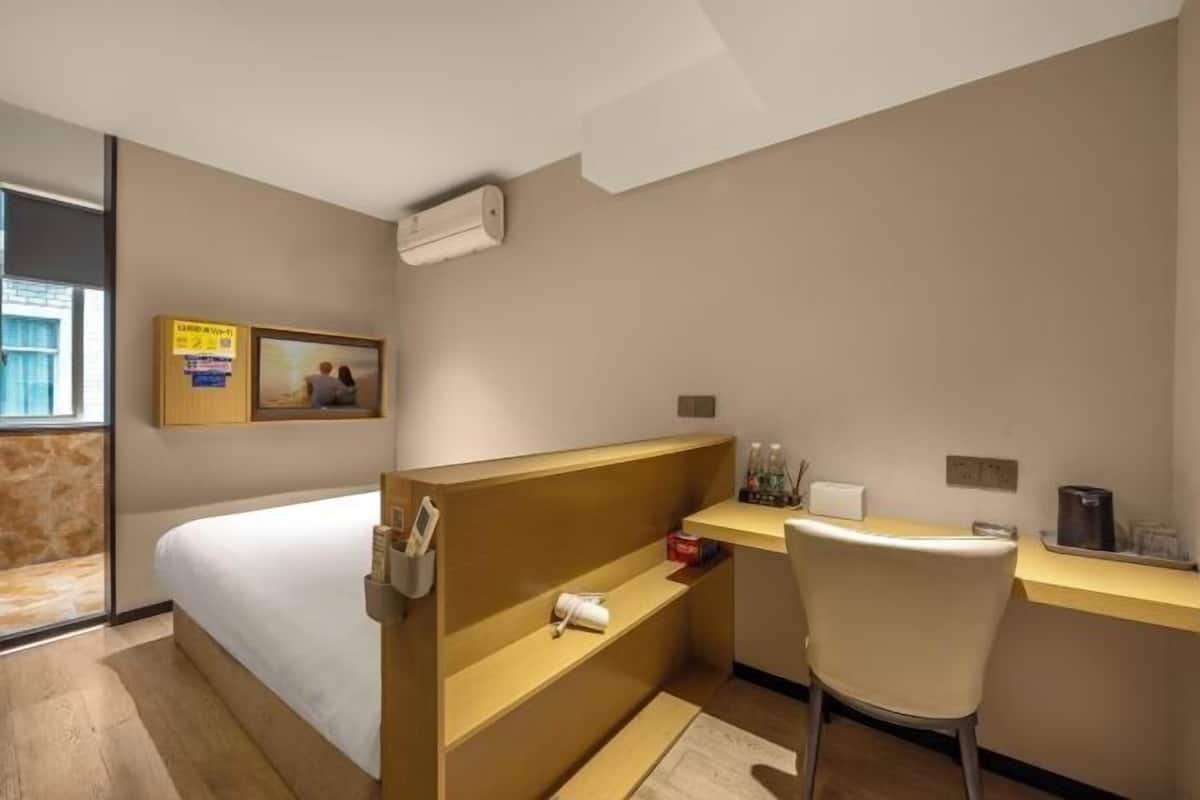 A hotel room with a bed, a desk, a chair, a shelf with toiletries, a picture on the wall, and a window.