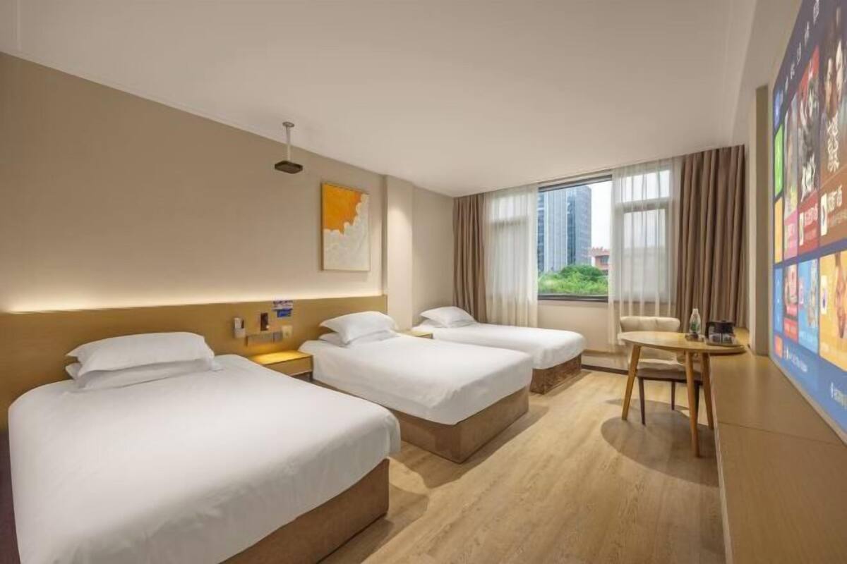 A hotel room with two beds, a small table, a chair, a window with curtains, and a wall-mounted TV.