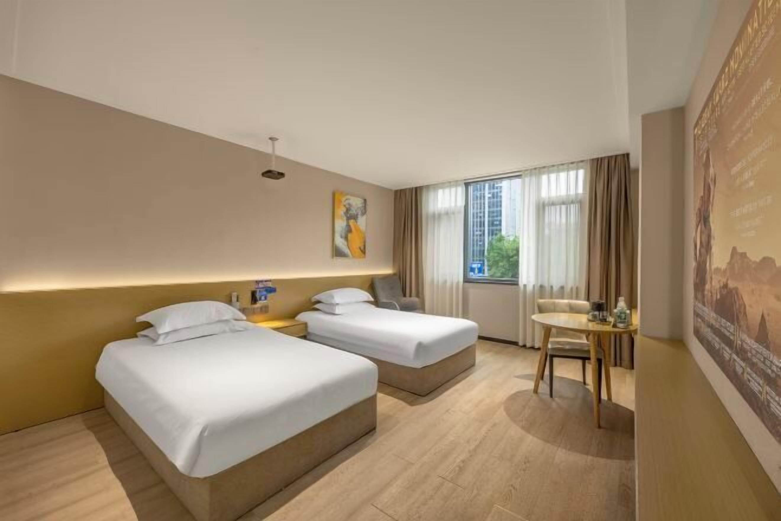 A hotel room with two single beds, a small table with a chair, a large window with curtains, and a wall-mounted artwork.