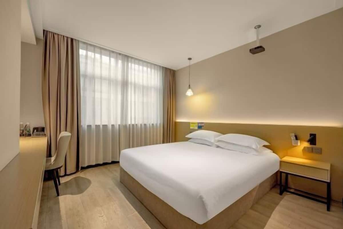 A hotel room with a bed, bedside table, lamp, curtains, and a desk with a chair.