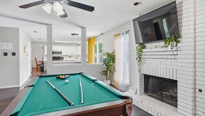A pool table in a room with a fireplace, a ceiling fan, and a view to a kitchen area.