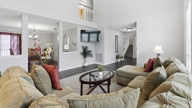 A spacious living area with a high ceiling, a flat-screen TV mounted on the wall, a round coffee table, and a staircase leading to an upper level.