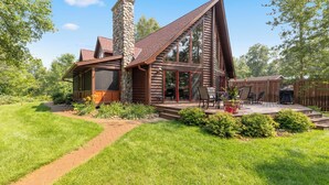 Une cabane en rondins avec une cheminée en pierre, de grandes fenêtres et une terrasse aménagée avec du mobilier d’extérieur.