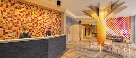 A hotel reception area with a patterned wall, a modern reception desk, and a seating area with chairs and a table.