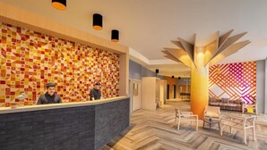 Un espace d’accueil d’hôtel avec un mur décoré d’un motif, un comptoir d’accueil moderne et un coin salon avec des chaises et une table.