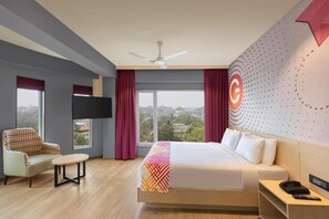 A hotel room with a bed, a chair, a small table, a ceiling fan, a TV, and a large window with curtains. - Ginger Kochi, Kalamassery (Kanayannur)