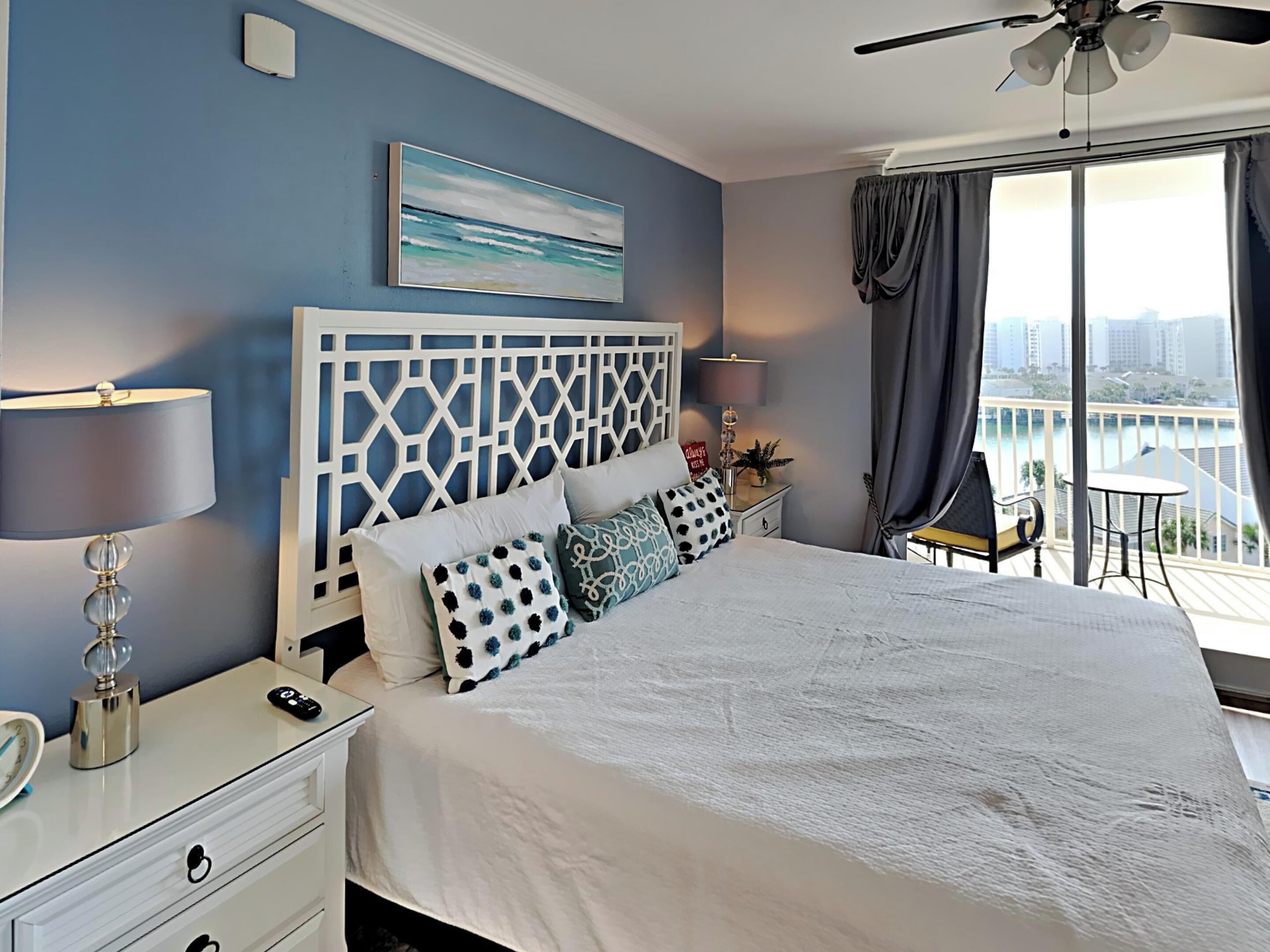 A bedroom with a bed, a nightstand, a lamp, a ceiling fan, and a view of the city through large windows.