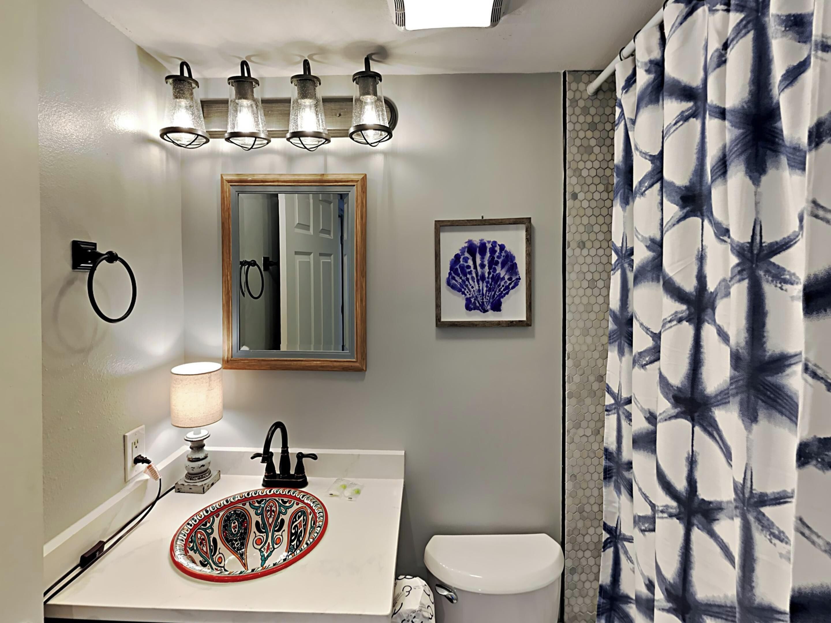 A bathroom with a white sink, black faucet, and a decorative plate. There are three wall-mounted lights, a framed mirror, and a toilet.